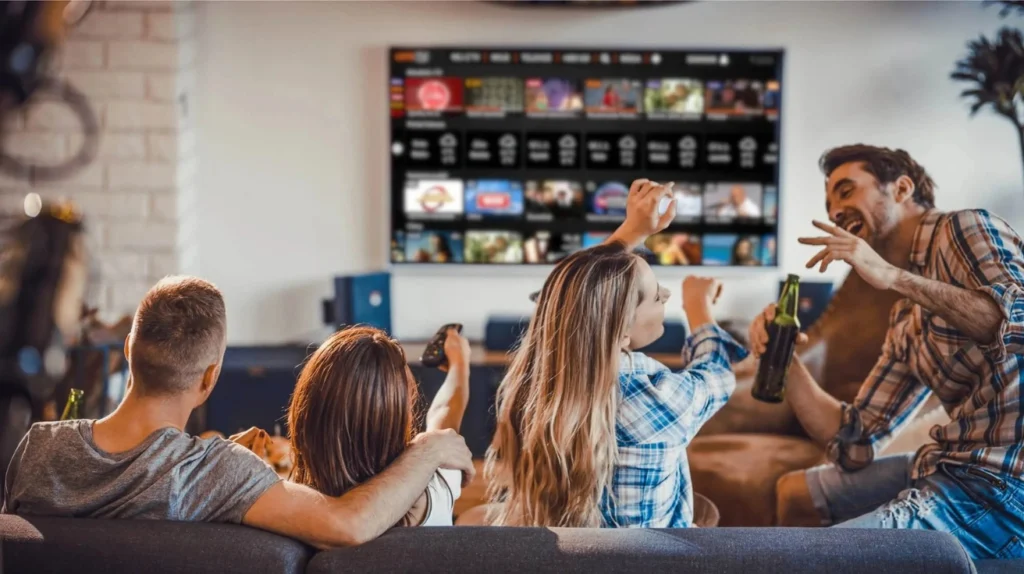 This image shows a group of four friends enjoying entertainment on a large TV. They're sitting on a couch facing a wall-mounted TV displaying what appears to be a streaming service interface with multiple video thumbnails arranged in a grid. The friends are celebrating or reacting enthusiastically to something on screen - one person is raising their arms in excitement, another is holding what looks like a bottle, and they all appear to be having a good time in a home setting with a white brick wall visible in the background.