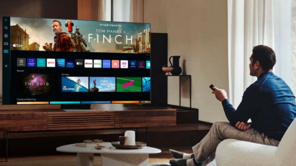 This image shows a man in a blue blazer sitting in what appears to be a living room, holding a remote control while watching a large TV. The television is displaying the Apple TV+ interface featuring the movie "Finch" starring Tom Hanks. The screen shows the film's promotional banner at the top with Tom Hanks in an orange jacket, and below it are rows of app icons including Netflix, Prime, and other streaming services, followed by content recommendations. The TV sits on a dark wooden media console, and there's a small white coffee table in the foreground with some items on it. The room has neutral-colored walls with some decorative elements visible in the background.