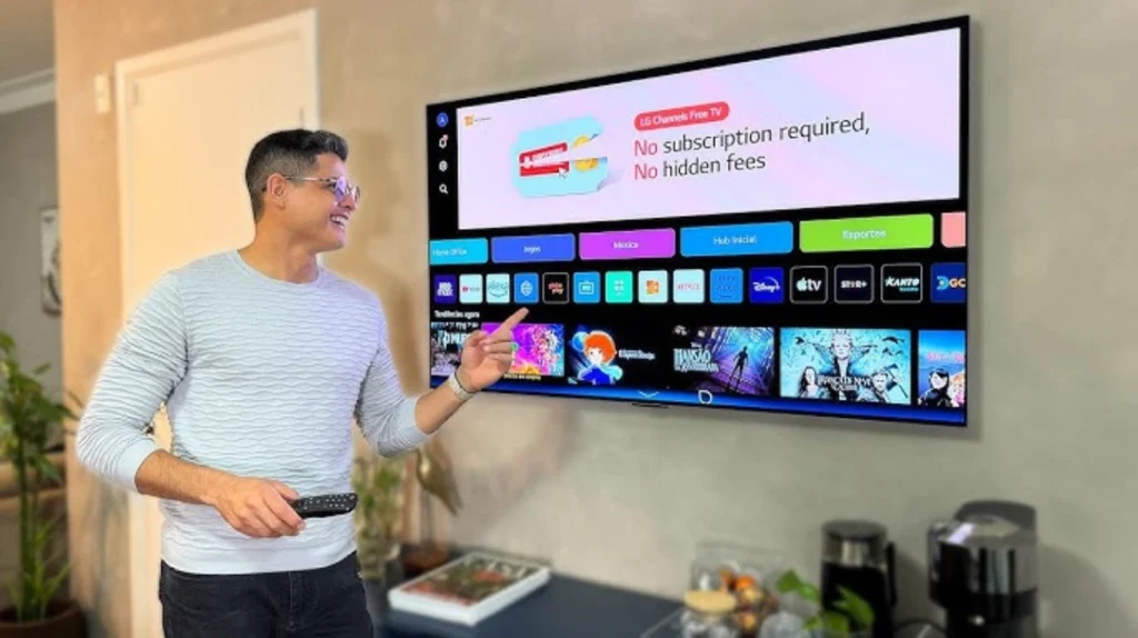 This image shows a man with glasses and short dark hair wearing a light gray/white striped long-sleeve shirt, standing next to a wall-mounted TV while holding a remote control. He's smiling and pointing at the TV screen, which displays an LG smart TV interface. The screen shows a banner for "LG Channels Free TV" with text stating "No subscription required, No hidden fees." Below this are rows of colorful app icons for various streaming services and a row of content thumbnails showing different shows or movies. The setting appears to be a home with a gray wall, and there's a countertop or table visible at the bottom of the image with some household items.
