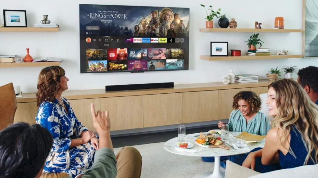 This image shows a group of people in a modern living room watching a television displaying the Amazon Prime Video interface. The TV is showing "The Lord of the Rings: The Rings of Power" promotional banner at the top, with streaming service icons visible below it including Netflix, Amazon Prime, and others. The room features floating wooden shelves with decorative items and plants, and a light wood media console beneath the TV with a soundbar. In the foreground, several people are gathered with some sitting on furniture while enjoying snacks and drinks at a small round table. A woman in a blue and white patterned dress appears to be speaking while another person is raising their hand.