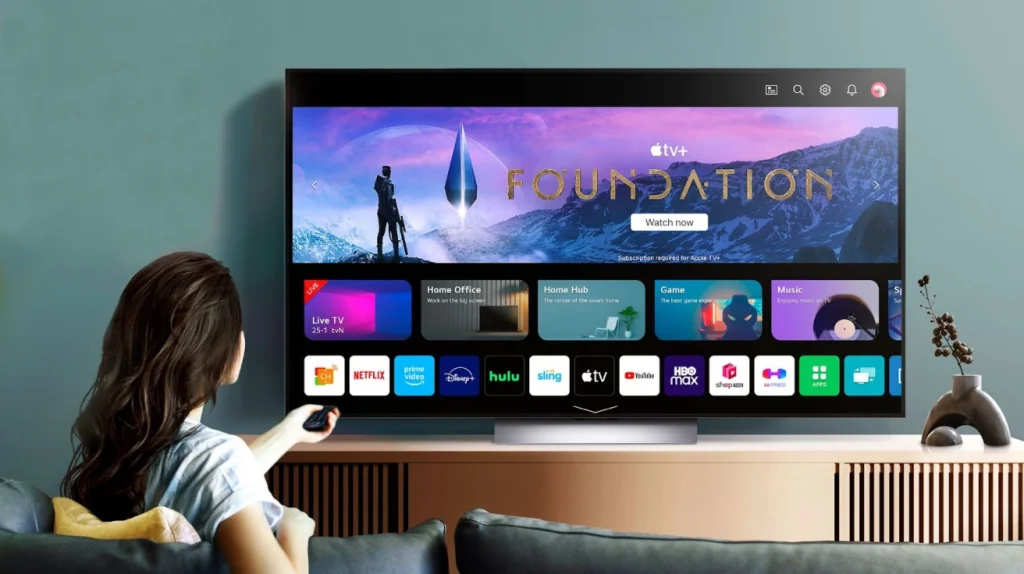 This image shows a person with long dark hair sitting on a couch, pointing a remote control at a large television. The TV screen displays the Apple TV interface featuring a banner for the Apple TV+ show "Foundation" with a "Watch now" button. Below the banner are various content categories like "Live TV," "Home Office," "Home Hub," "Game," and "Music." The bottom row shows app icons for streaming services including Netflix, Prime Video, Disney+, Hulu, Sling, Apple TV, YouTube, HBO Max, and others. The TV is mounted on a teal/blue wall and sits on a wooden media console with a small decorative item visible on the right side.