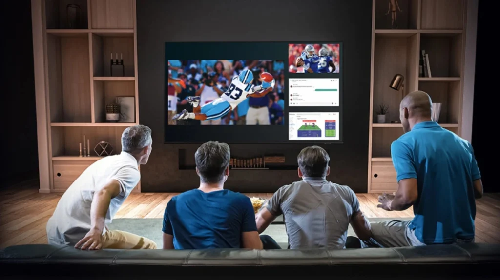 This image shows four men sitting on a dark leather couch watching football/American football on a large TV. They are viewing from behind, facing a wall-mounted TV that displays a football game featuring a player in a blue jersey (number 23) making a diving catch or play. The TV screen also shows additional content on the right side, including what appears to be stats or chat features. The men are in a modern living room with wooden built-in shelving units on either side of the TV. They appear engaged in watching the sports content, with one man in a turquoise shirt on the right side of the couch.RetryClaude can make mistakes. Please double-check responses.